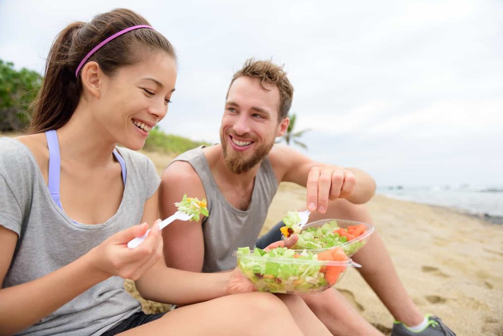 A smiling man and woman sit on a sandy beach, dressed in casual workout clothes, sharing a healthy meal from clear plastic bowls. The woman has a headband and laughs while holding a forkful of salad. The man, with a beard, looks at her while holding another bowl of salad.