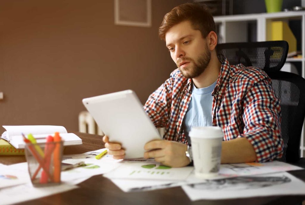 A man with a beard, wearing a red checkered shirt over a blue t-shirt, is seated at a desk holding a tablet. The desk is cluttered with papers, a coffee cup, and colorful markers in a holder. Shelves are visible in the background.