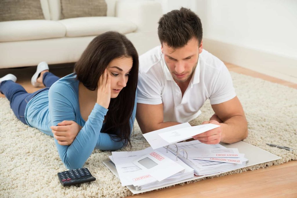 A couple lies on the floor of a living room with a white couch in the background. They are surrounded by bills and documents, with the woman looking concerned and holding her head while the man reads a letter. A calculator rests nearby.