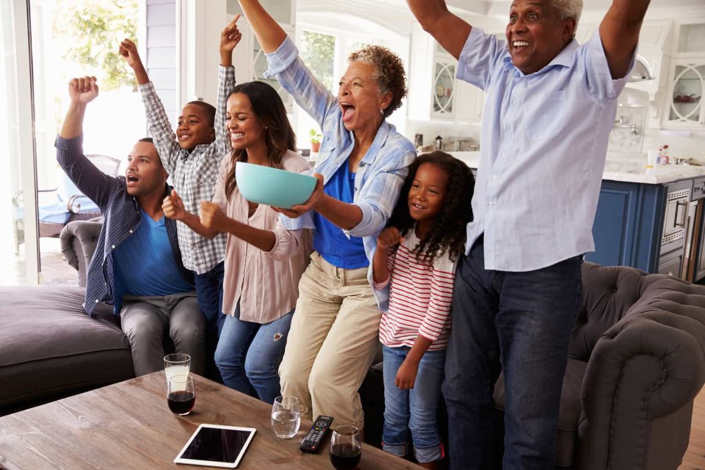A diverse family of six, including two elderly individuals, two adults, and two children, are sitting on a couch and cheering enthusiastically. They are watching something exciting on TV in a brightly lit living room with a cozy atmosphere.