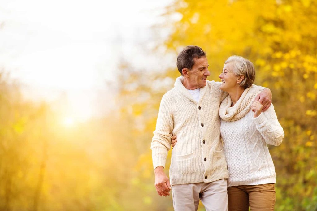 An elderly couple, both wearing cozy sweaters, walk arm in arm through a sunlit park with autumn foliage. They are smiling at each other, creating a warm and joyful atmosphere.