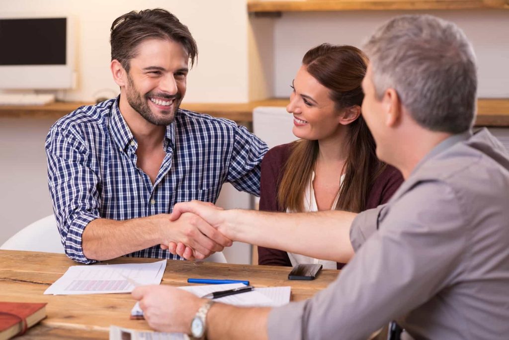A man and woman smile while sitting at a table and shaking hands with another man. Papers and a pen are on the table.