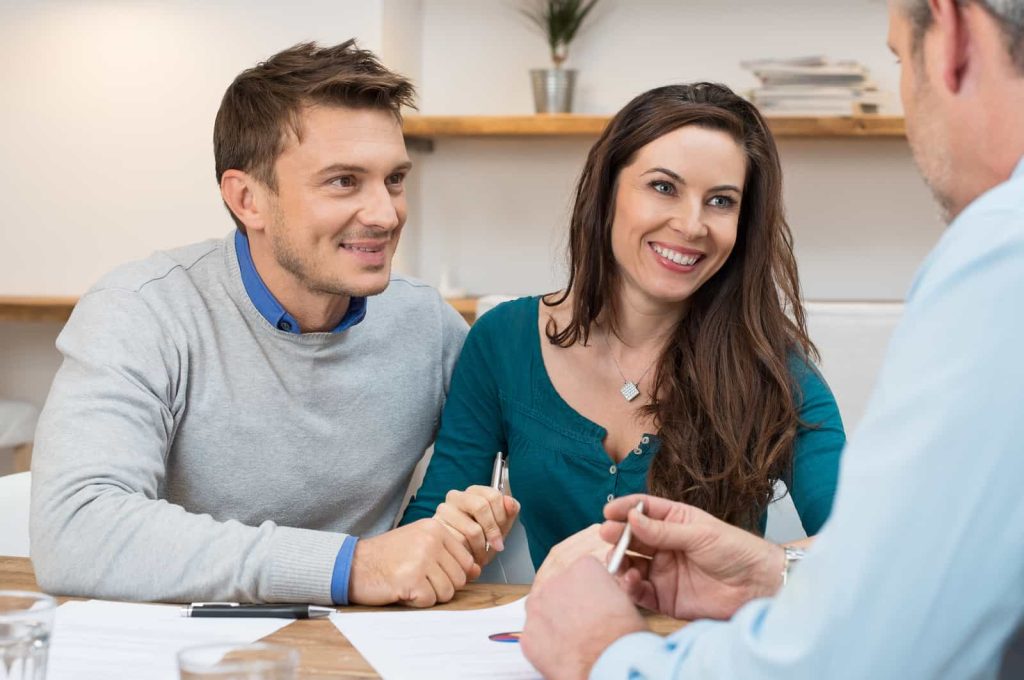A smiling couple sits at a table across from a man in a light blue shirt. They are engaged in a discussion and hold pens, possibly preparing to sign documents.