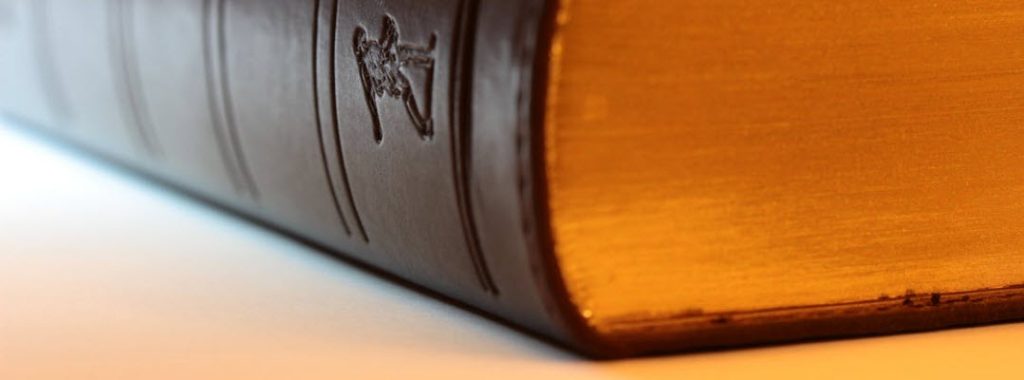 A close-up shot of a large, thick book with a dark brown cover and gold-edged pages. The textured spine and gilded pages give it a vintage and elegant appearance.