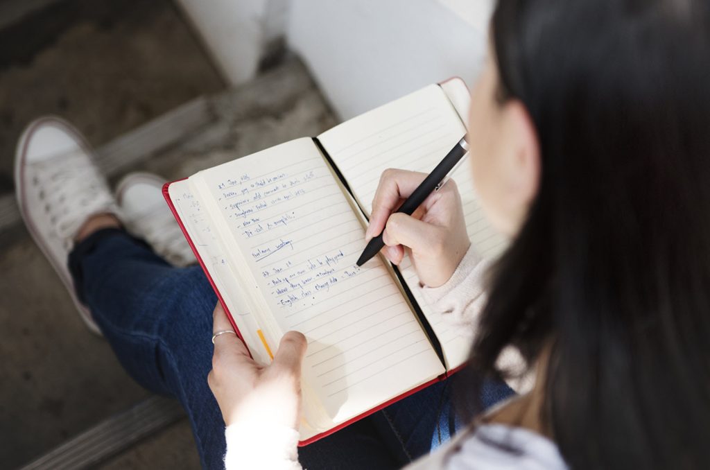 A person with long dark hair, wearing a light-colored sweater and blue jeans, is sitting on steps and writing in an open notebook with lined pages. The person holds a pen in their right hand and appears to be making notes or a journal entry.