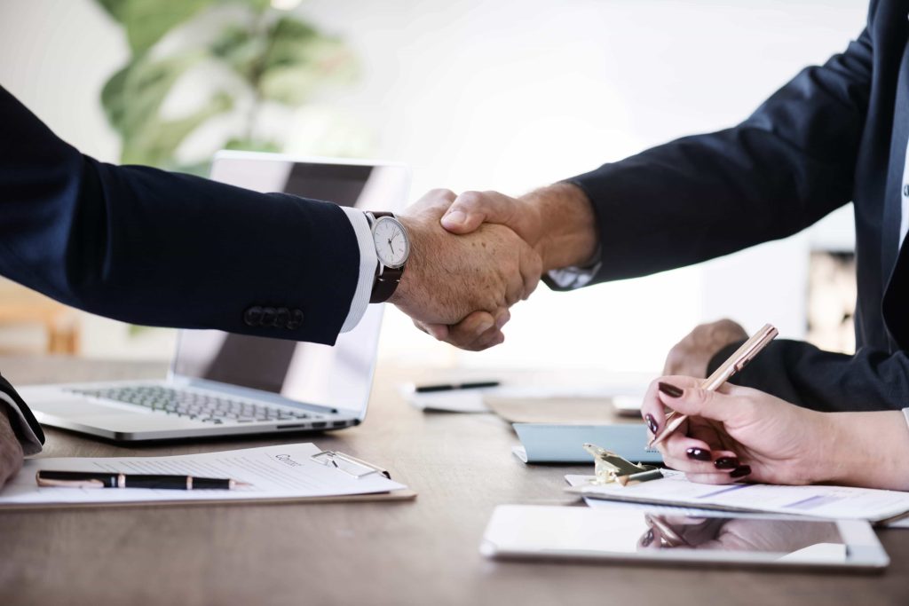 Two people in business attire shaking hands over a wooden desk with documents, a laptop, and a tablet. Another person is taking notes beside them with a pen and notepad.