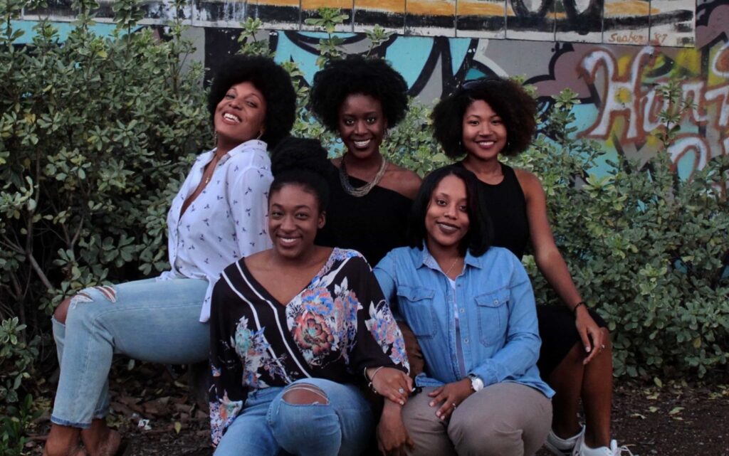 Five women posing together outdoors in front of a graffitied wall. They are smiling and casually dressed, sitting and standing among green bushes. The mood is joyful and relaxed, capturing a moment of friendship and camaraderie.