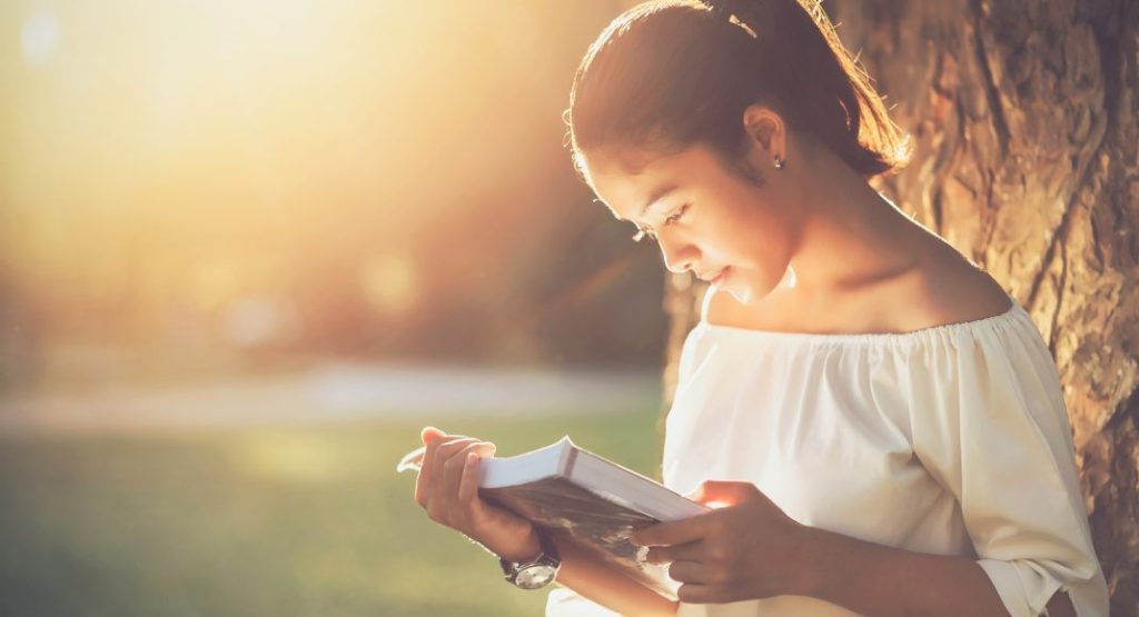 A young woman with her hair tied back is leaning against a tree while reading a book. She is wearing an off-shoulder white top and is illuminated by warm sunlight, creating a serene and peaceful atmosphere.