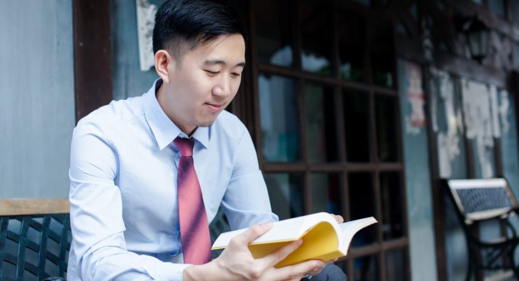 A man in a light blue dress shirt and red tie sits outside on a metal bench, reading a yellow book. He appears focused and content.