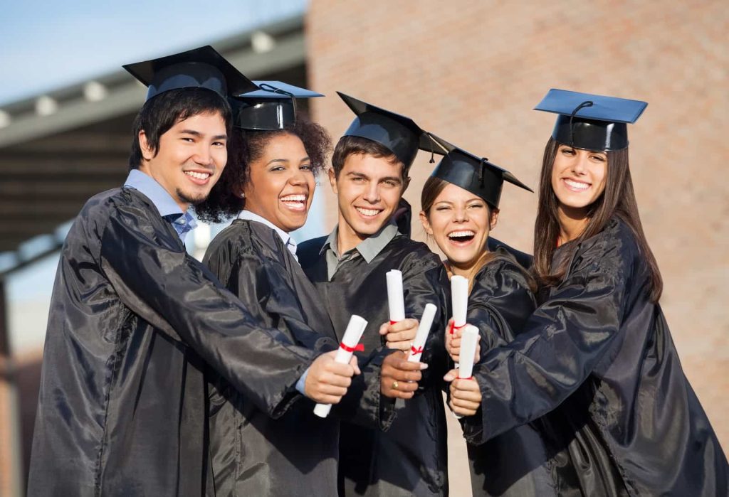 Five graduates dressed in black graduation caps and gowns stand closely together, smiling and holding diplomas towards the camera. They appear joyful and excited.