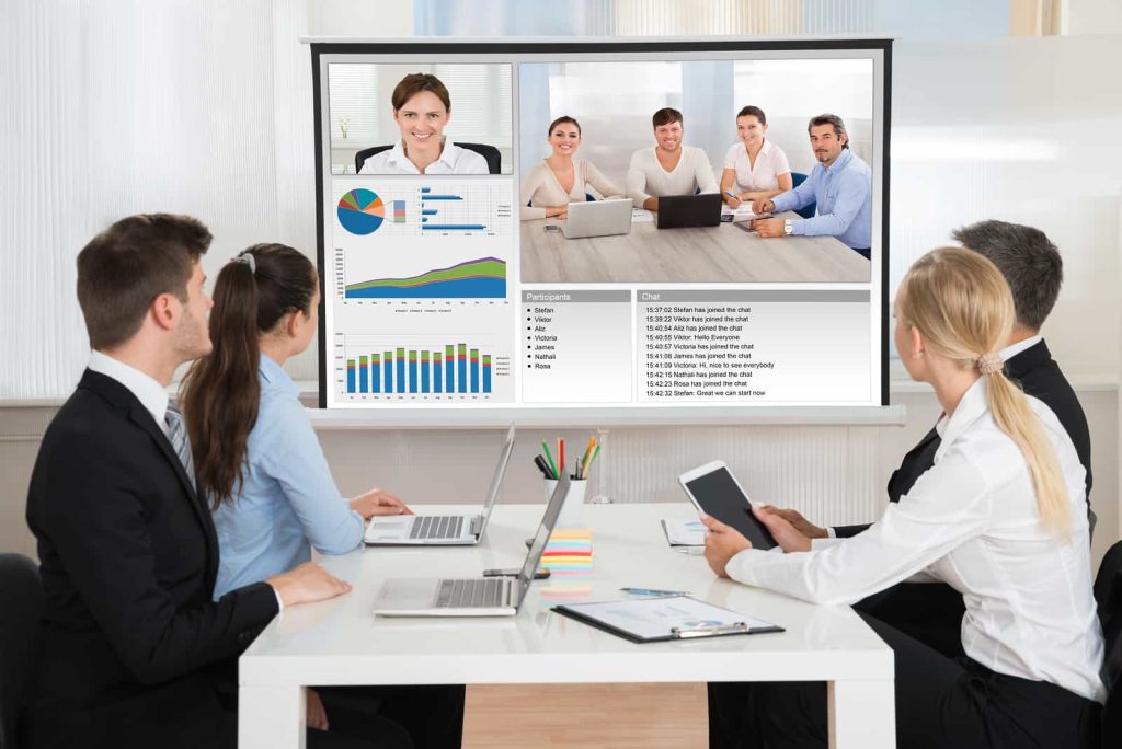 Five people sit around a table with laptops and tablets, attentively watching a screen displaying a video conference. The screen shows participants, charts, and a data table.