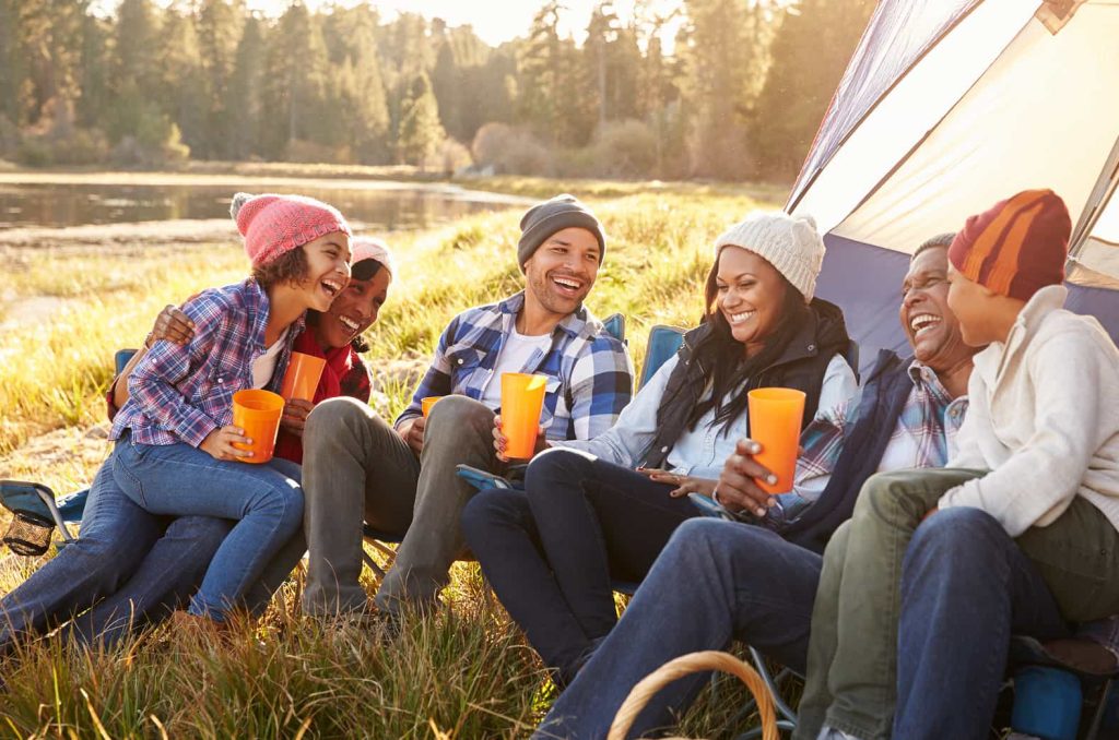 A group of six people, dressed casually in warm clothing and hats, are sitting on camping chairs outside a tent by a lake. They are smiling and laughing while holding orange cups, enjoying a relaxing moment in a scenic, wooded area during the day.