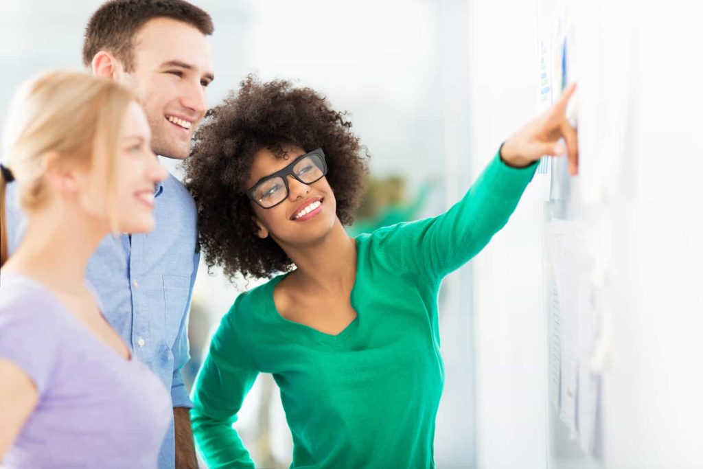 Three people are smiling and looking at a bulletin board. The person in the foreground, wearing glasses and a green shirt, is pointing at something on the board. The other two, a woman in a purple shirt and a man in a blue shirt, look at the bulletin board.