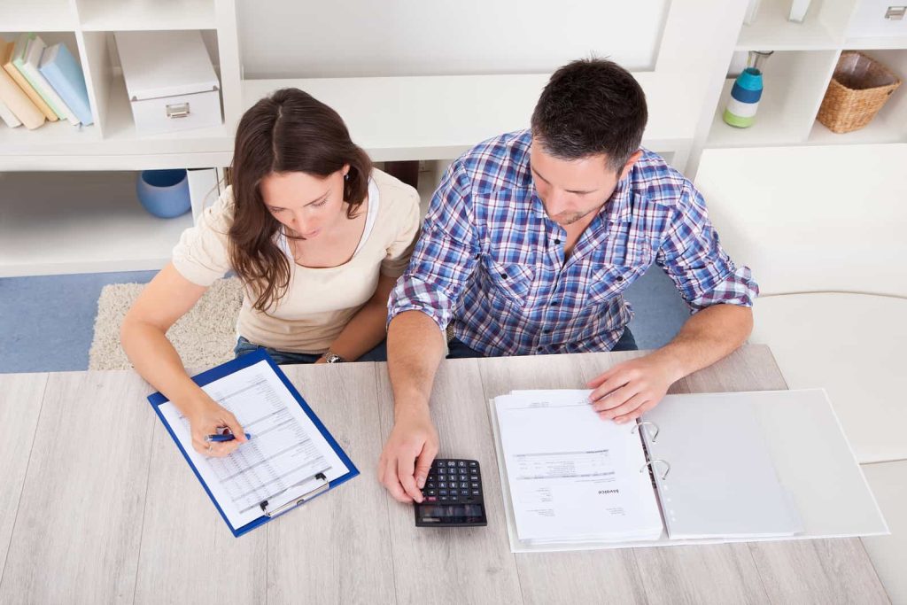 A man and woman sit at a table working together. The woman is reviewing documents on a clipboard and writing with a pen. The man is using a calculator and looking at papers in a binder. Shelves with folders and decorative items are in the background.
