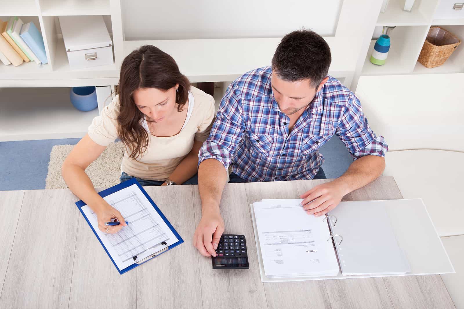 A man and woman sit at a table working together. The woman is reviewing documents on a clipboard and writing with a pen. The man is using a calculator and looking at papers in a binder. Shelves with folders and decorative items are in the background.