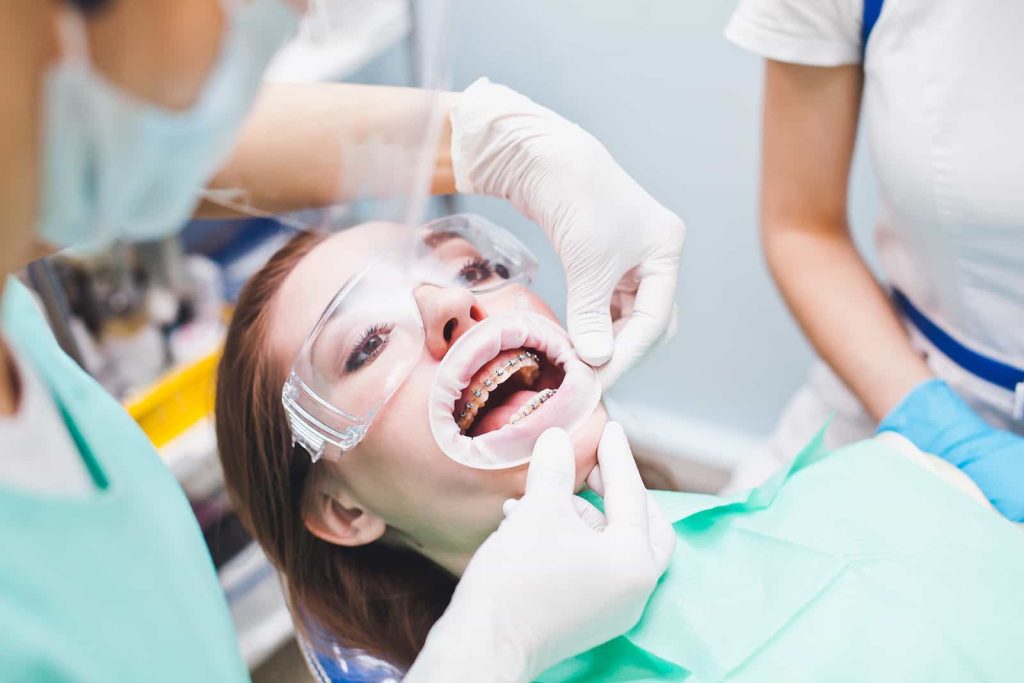 A woman wearing protective glasses is having her teeth examined at a dental clinic. She has braces and a mouth retractor in place, while two dental professionals, both wearing masks and gloves, are working on her teeth.