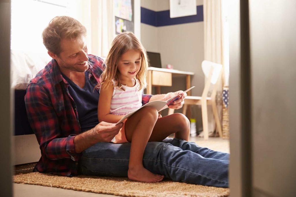 A father and a young daughter are sitting on the floor in a cozy room. The father is reading a book to the girl, who is sitting on his lap and looking at the pages intently. They both have smiles on their faces, and the room is warmly lit with simple decor.