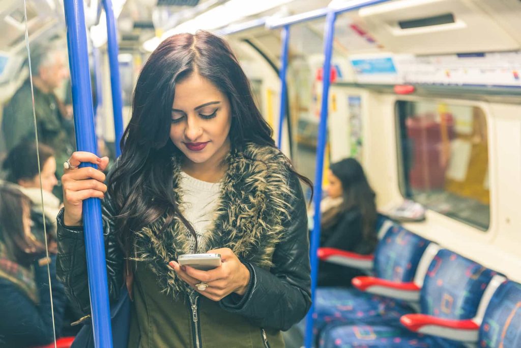 A woman with long dark hair and a fur-lined jacket stands in a subway car, holding onto a blue pole with one hand and looking at her smartphone with the other. Passengers are seated in the background.