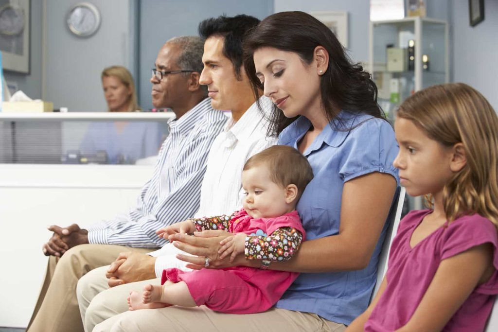 A diverse group of people, including an older man, a middle-aged man, a woman holding a baby, and a young girl, are sitting in a waiting room. The room has a counter and some medical equipment in the background.