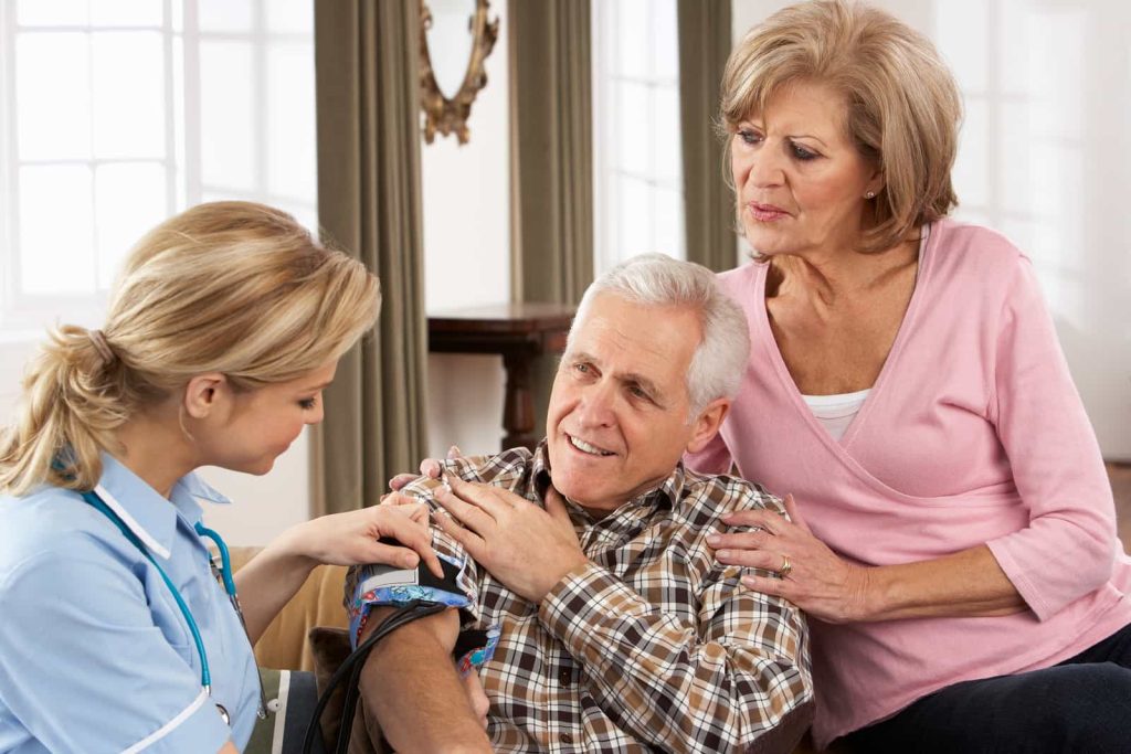 A nurse checks the blood pressure of an elderly man seated on a couch, as an elderly woman looks on with concern in a well-lit living room.