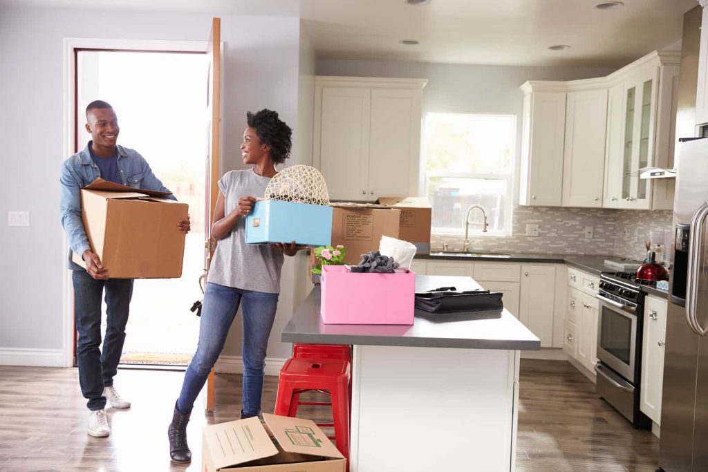 A man and a woman are in a bright, modern kitchen. The woman is holding a box and a basket with kitchen items. The man follows behind carrying two cardboard boxes. More moving boxes are placed on the floor and kitchen counter.