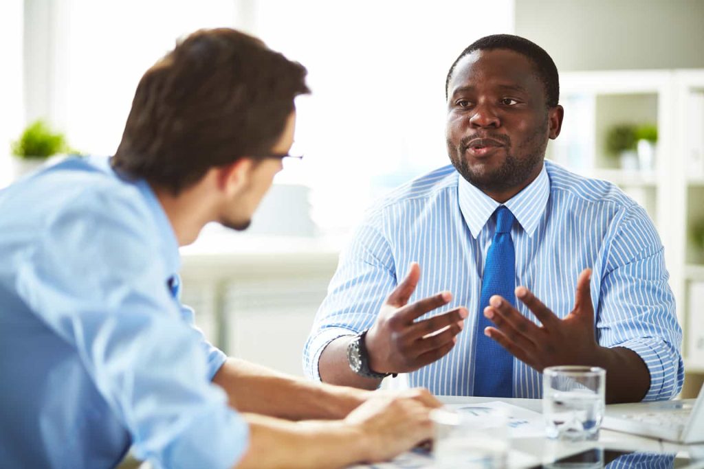 Two businessmen in conversation at a table in a bright office setting, one gesturing while speaking and the other listening attentively.