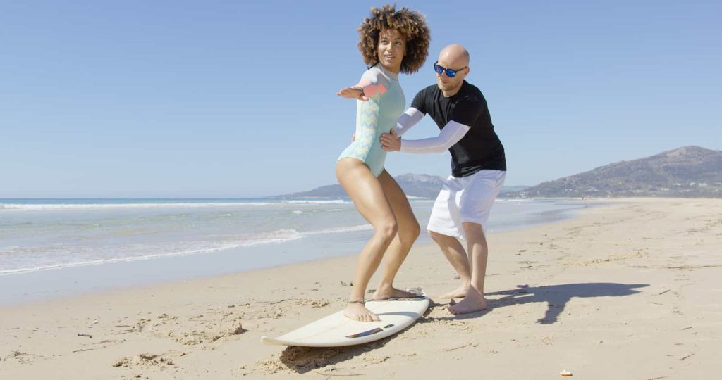 A man teaches a woman how to surf on a sandy beach on a sunny day.