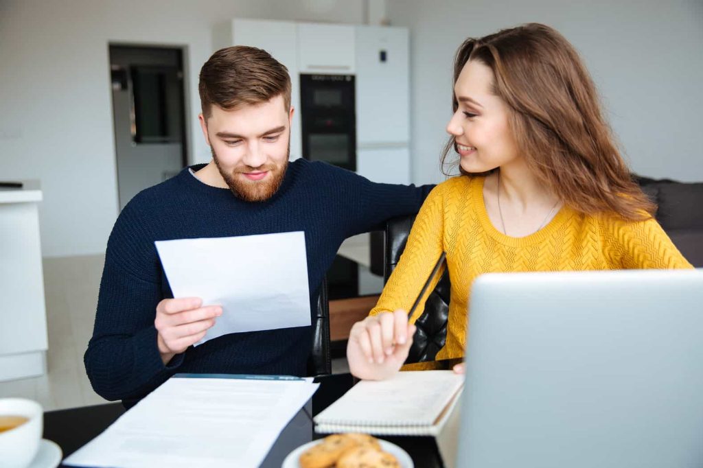 A young couple reviews documents together at a kitchen table, with a laptop open and snacks nearby, in a well-lit, modern kitchen.