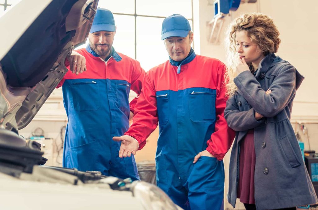 Two mechanics in blue and red coveralls consult with a female client, showing her something under the hood of a car in a bright garage.