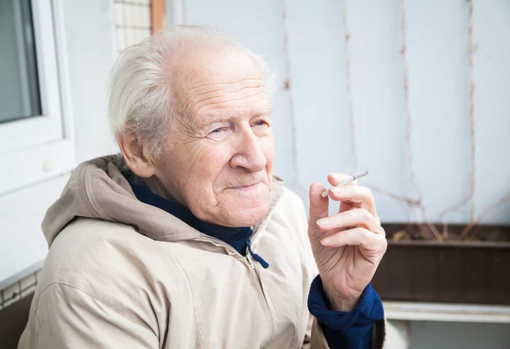 An elderly man with grey hair sits outside, wearing a beige jacket and a blue shirt, smiling slightly as he holds a lit cigarette.
