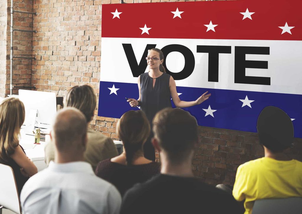 A woman presents to an attentive audience in front of a large "vote" banner with a star-spangled design, in a room with brick walls and bright lighting.