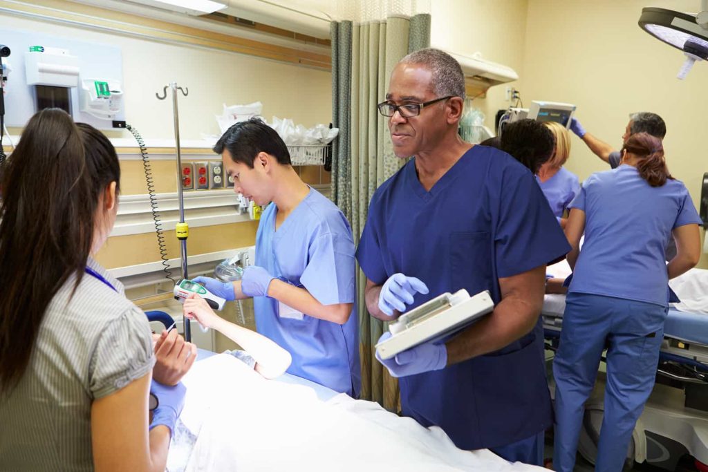 A diverse medical team working in a busy hospital room, with a senior male doctor holding a clipboard and discussing with a patient. In the background, other medical staff assist.