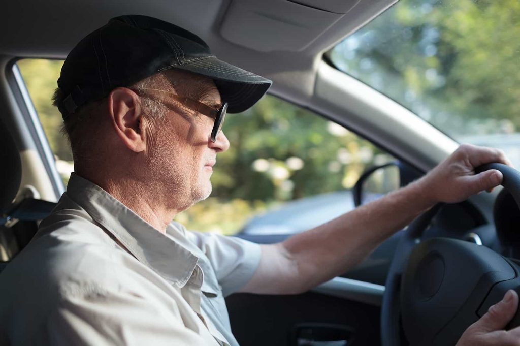 Senior man with a cap driving a car, focusing on the road with sunlight streaming through the window.