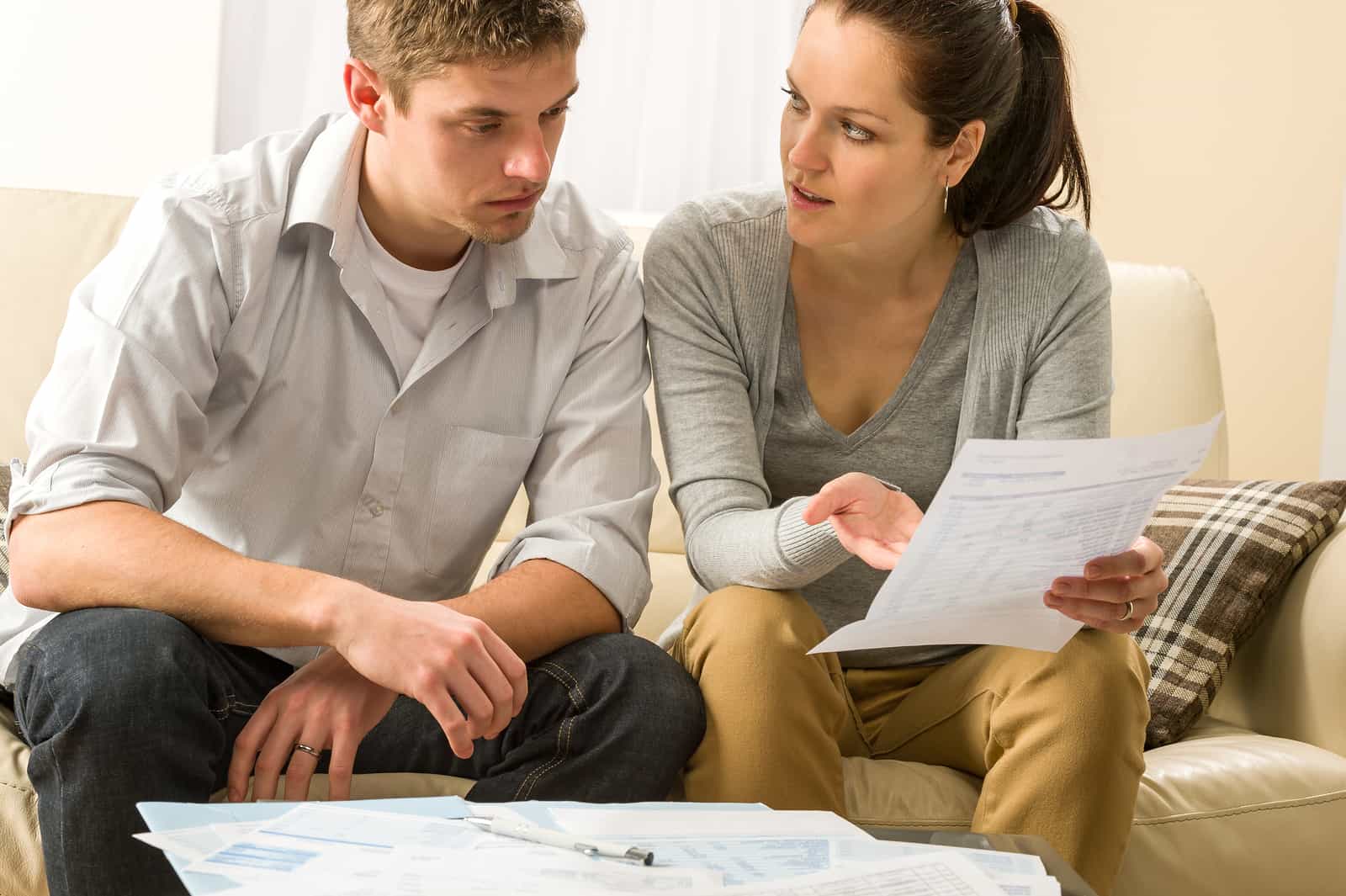 A young couple sits on a cream-colored couch, looking concerned as they review documents and bills spread out on the coffee table in front of them.