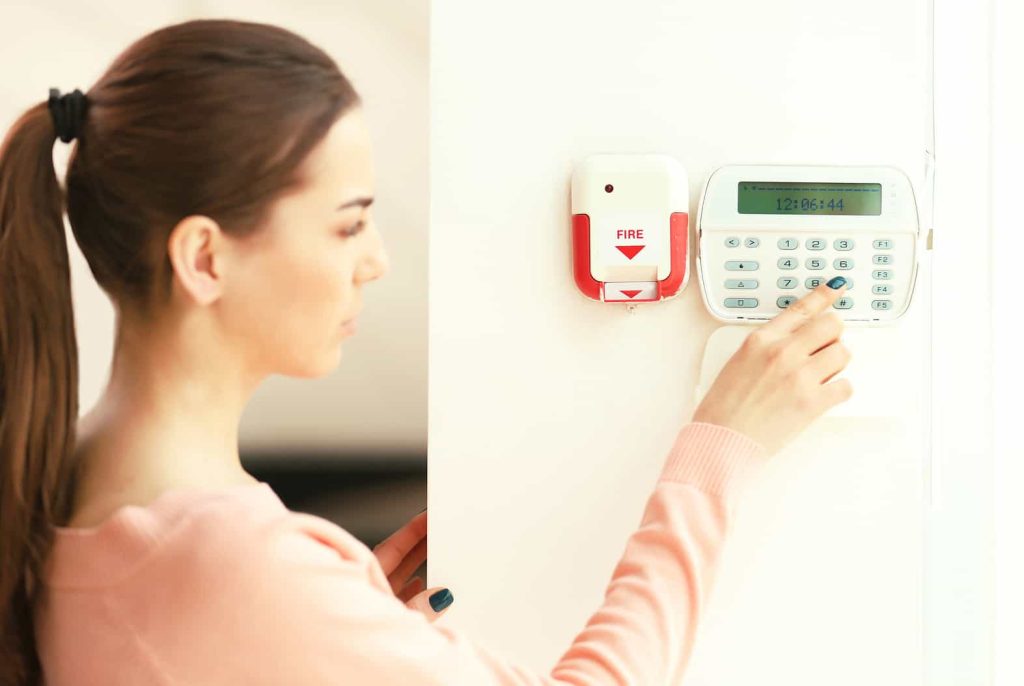 A woman in a pink sweater is programming a security keypad next to a red fire alarm on a white wall.