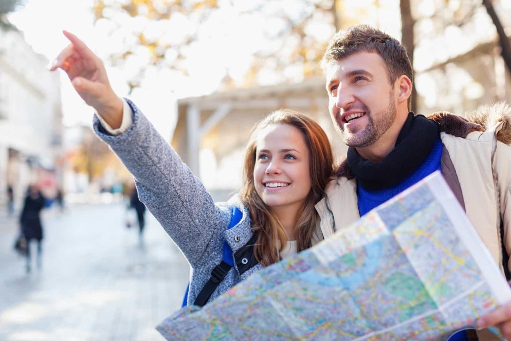 A joyful young couple with a map exploring a city street on a sunny day, with the woman pointing at something interesting while the man looks in the direction she indicates.