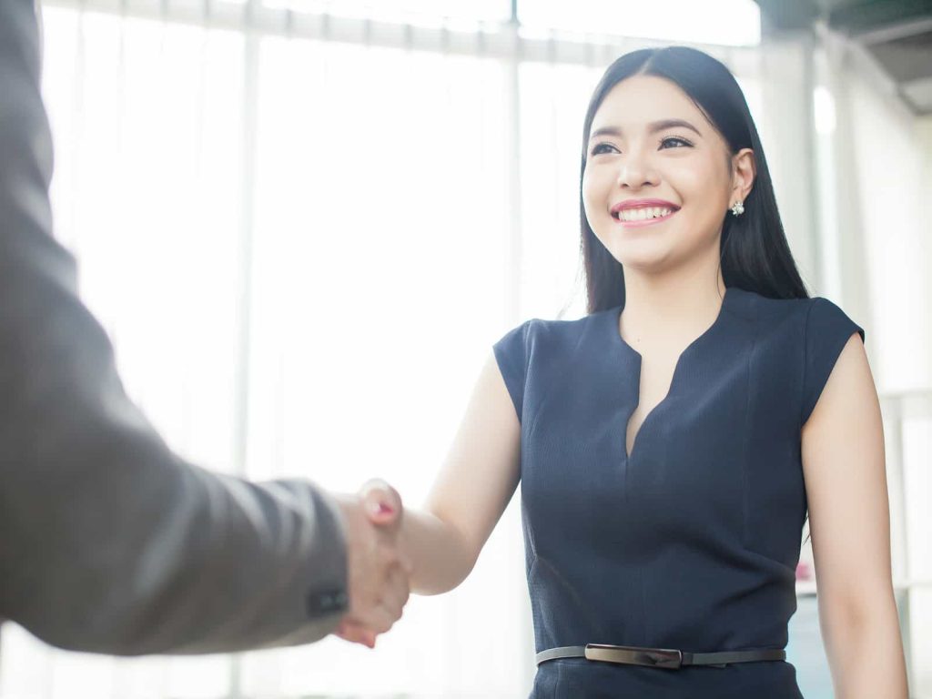 A professional woman in a business outfit smiling and shaking hands with someone, in a bright office environment.