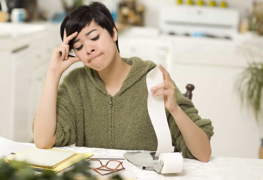 A woman with short hair sits at a table, looking stressed as she examines a long receipt from a calculator beside notebooks and glasses.