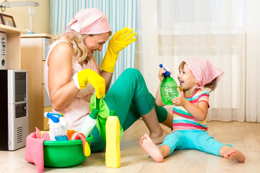 A woman and a young girl in matching headscarves and gloves playfully engage in house cleaning, sitting on a kitchen floor surrounded by cleaning supplies.