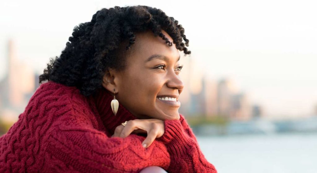 A joyful young woman with curly hair, wearing a red sweater and earrings, smiling and looking away against a blurred cityscape background during sunset.