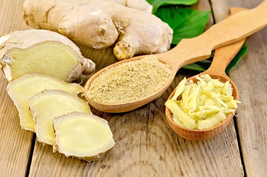 Whole and sliced ginger root with a wooden spoon of ginger powder and another spoon of grated ginger, on a rustic wooden table.