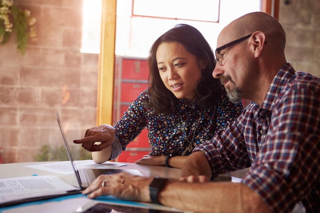 A man and a woman collaborate at a desk, discussing over a laptop and documents in a brightly lit office space with a rustic brick wall in the background.