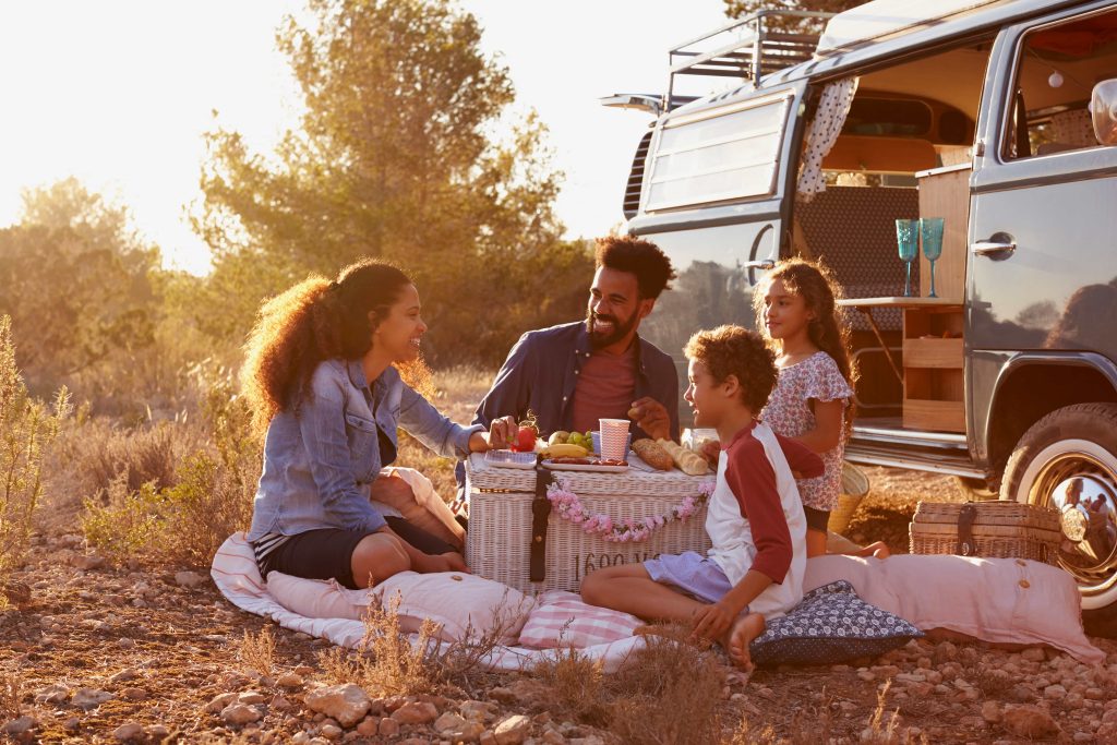 A family enjoys a picnic beside their camper van in a sunlit natural setting, sharing food and laughter on a comfy blanket with natural scenery around.