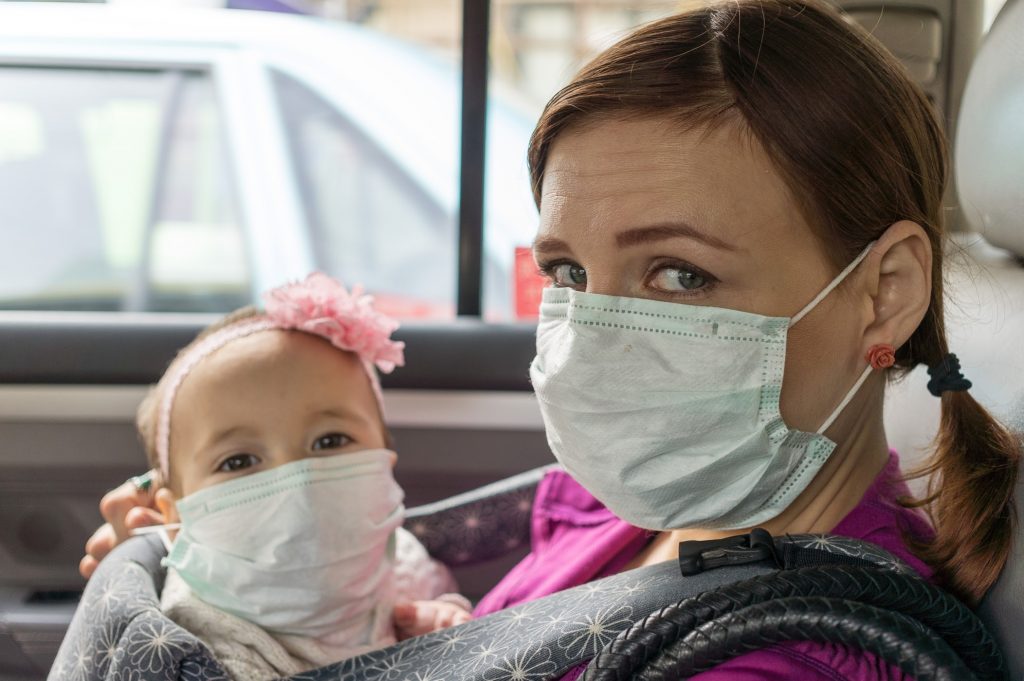 A woman wearing a medical mask holds a baby, also in a mask, inside a vehicle. They are looking towards the camera, the woman with a subtle smile.