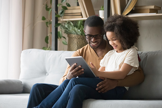 A father and a daughter, both smiling, sit on a sofa while looking at a tablet they are holding together, in a cozy room with books and plants in the background.