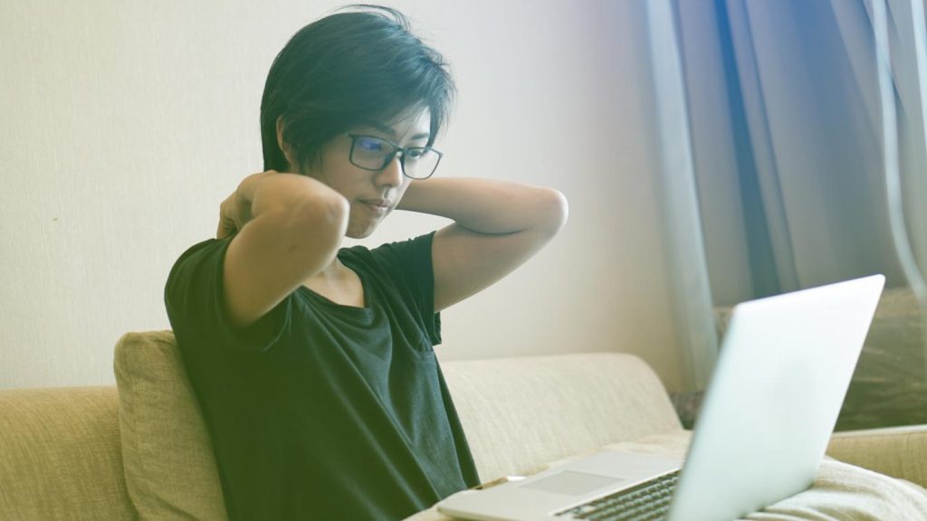A person with glasses sitting on a sofa, working on a laptop, and stretching their neck. Natural light fills the room from a window in the background.