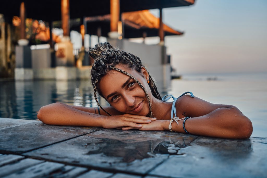 A woman with braided hair smiles while resting her chin on her hands at the edge of a swimming pool, with a blurry background of a resort during sunset.