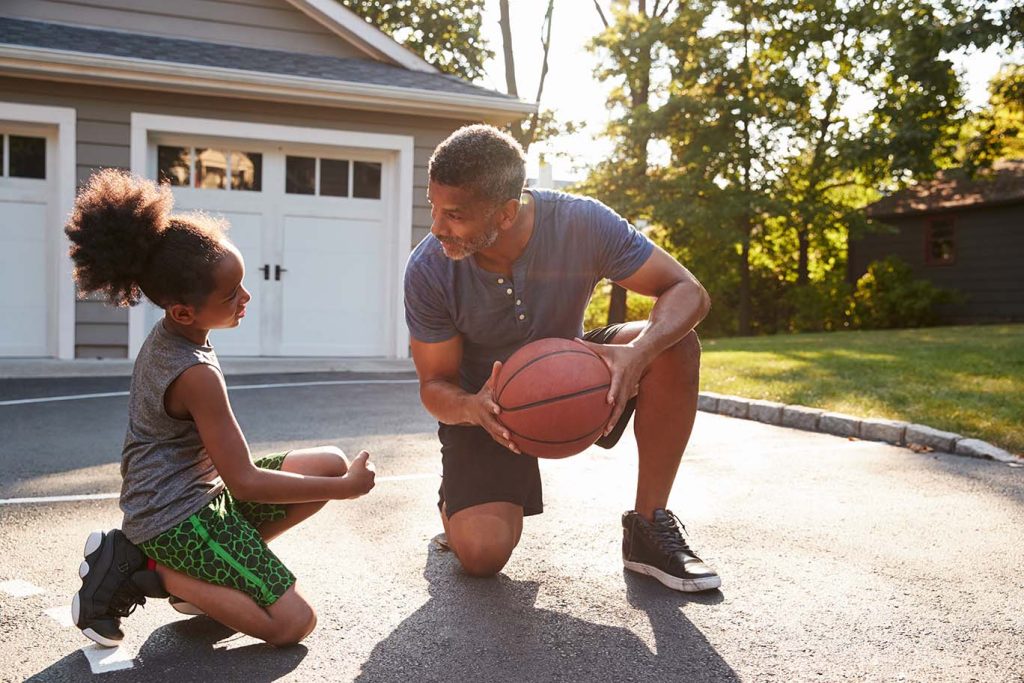 A father and a young daughter playing basketball together in a driveway, with a house and garage in the background, during a sunny day.