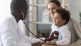 A doctor using a stethoscope on a young boy, who is smiling and sitting with his mother in a medical office.