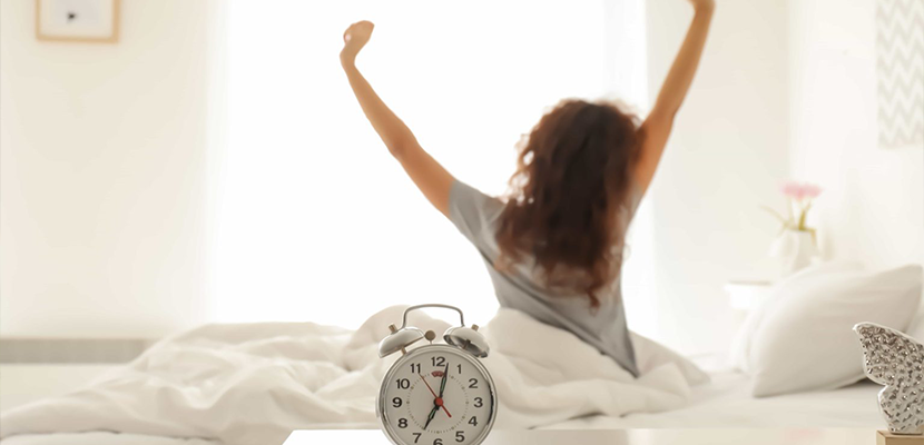 A woman sits on a bed, stretching her arms upwards with a feeling of refreshment. a classic alarm clock shows the time in a warmly lit bedroom.